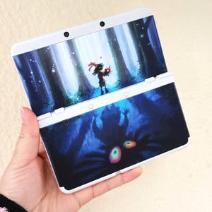 Puede incluir: Consola de juegos portátil Nintendo 3DS blanca con una cubierta personalizada. La parte superior muestra una escena de bosque con un personaje sosteniendo objetos brillantes. La parte inferior muestra una criatura azul oscuro con ojos brillantes.