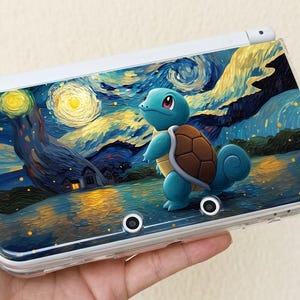 Op de afbeelding: Een draagbare spelcomputer met een beschermende skin voorzien van een levendige, schilderachtige illustratie van een blauwe schildpad bij een waterkant onder een wervelende, sterrenhemel met heldere gele bollen en diepblauwe tinten.