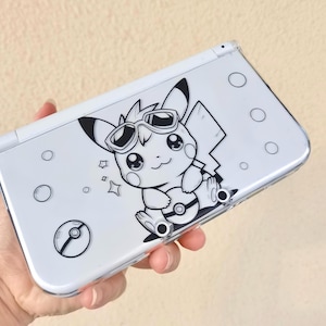 Könnte beinhalten: Eine weiße Nintendo 3DS Handheld-Spielkonsole mit einer transparenten Schutzhülle. Die Hülle zeigt eine schwarz-weiße Grafik von Pikachu mit Sonnenbrille und einem Pokéball. Die Konsole hat mehrere schwarze Kreise und ein Pokéball-Design.