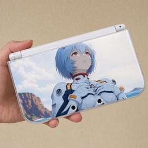 May include: A white handheld gaming console featuring a clear protective cover with a printed graphic of an anime character with short blue hair and red eyes, wearing a white and blue mechanical suit against a coastal landscape background.