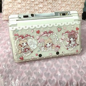 Può includere: Una custodia bianca per console di gioco portatile con una sovrapposizione trasparente con un simpatico design di ragazza gatto. Il design include fiocchi rosa, fragole e cuori. La custodia è su una superficie di pluriball rosa.