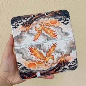 May include: A handheld gaming console case featuring a detailed illustration of a phoenix flying amidst volcanic mountains, smoke, and a glowing sun. The artwork displays vibrant orange and yellow flames against a backdrop of grey clouds and dark terrain.