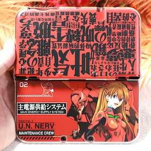 May include: Red and black handheld gaming console case with Japanese text and anime illustrations. The case is hinged and features the text "MAIN ENERGY SUPPLY SYSTEM" and "U.N. NERV MAINTENANCE CREW".