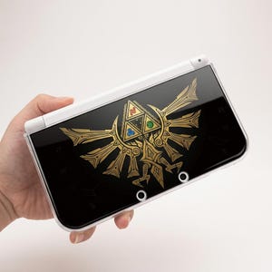 Può includere: Una console di gioco portatile bianca con una cover superiore nera decorata con un grande e intricato emblema color oro. L'emblema presenta un design triangolare centrale con piccole icone circolari rosse, blu e verdi integrate.