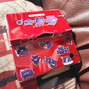 May include: A red Nintendo DS Lite handheld gaming console with a clear protective case. The case and console are decorated with various purple cartoon ghost stickers. The console is open, displaying the screen and buttons.