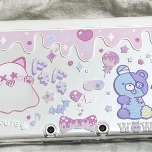 Pode incluir: Uma capa branca para Nintendo 3DS com um design rosa e roxo. A capa apresenta gráficos de desenhos animados de um fantasma, um urso e outros elementos fofos. O design inclui corações, estrelas e as palavras "CUTE" e "WOW".