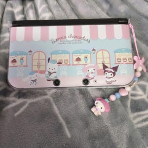 May include: A pink and blue handheld gaming console featuring Sanrio characters. The design includes a bakery storefront with various characters. A pink lanyard with a charm is attached to the console.