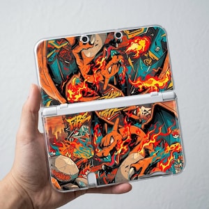 May include: A handheld gaming console featuring a vibrant graphic skin of a fire-breathing dragon character surrounded by flames, stylized graffiti art, and small circular icons. The design incorporates bright orange, red, and teal colors.