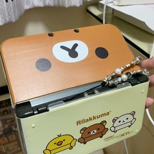 Könnte beinhalten: Eine hellbraune Nintendo 3DS LL-Spielkonsole mit Rilakkuma-Charakteren. Die obere Abdeckung hat ein Bärengesicht-Design. Die untere Hälfte ist hellgelb mit Rilakkuma und Freunden. Der Text "Rilakkuma" und "Nintendo 3DS LL" sind sichtbar.