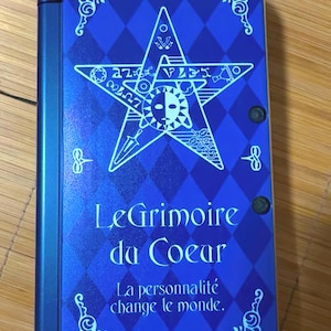 Puede incluir: Un libro azul con un patrón de diamantes y el texto "Le Grimoire du Coeur" y "La personalidad change le monde." Una estrella blanca con símbolos y un diseño de sol y luna está en el centro. El libro tiene una lomo azul y esquinas decorativas.