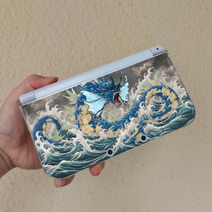 May include: A light blue handheld gaming console featuring a detailed graphic of a blue serpentine sea creature with a wide open mouth, surrounded by stylized white and dark blue ocean waves on a dark, cloudy background across the outer shell.