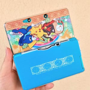 Può includere: Una console di gioco portatile Nintendo 3DS blu con una cover decorativa con i personaggi dei cartoni animati Pikachu, Ashimari, Nyabby e Mokuroh. La cover ha un design colorato con i nomi dei personaggi. La parte inferiore della console ha un design blu con il testo "©Nintendo Creatures GAME FREAK TV Tokyo ShoPro JR Kikako Pokémon."