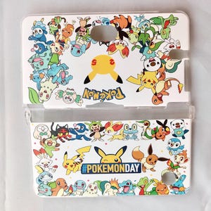 May include: White Nintendo DS case featuring a colorful array of Pokemon characters. The case includes a yellow Pikachu design and the text "Pokemon" and "#PokemonDay". The case is designed to protect the Nintendo DS.