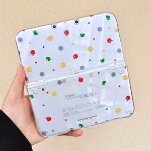 Puede incluir: Una consola de juegos portátil Nintendo 3DS LL blanca con un diseño de formas rojas, verdes, amarillas y grises. La consola está abierta, mostrando la pantalla y los botones. El texto "new: Nintendo 3DS LL" es visible.