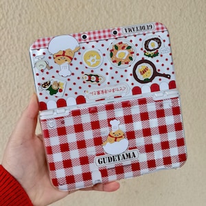 以下が含まれることがあります： 赤と白のチェック柄のニンテンドー3DSケース。グデタマのグラフィックが特徴です。ケースの下半分は赤と白のギンガムチェック柄、上半分はドット柄です。上半分には、さまざまなグデタマの食べ物のイラストが描かれています。