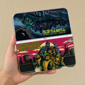 May include: A handheld gaming console featuring a Teenage Mutant Ninja Turtles graphic. The top screen shows a dark city scene with a crescent moon, while the bottom screen displays the four turtles and Shredder against a vibrant pink sky.
