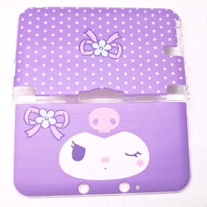 Puede incluir: Una funda morada para Nintendo 3DS con un estampado de lunares blancos en la mitad superior. La mitad inferior presenta una cara de personaje de dibujos animados con una calavera, un lazo y una estrella. La funda es de plástico y tiene una bisagra de plástico transparente.