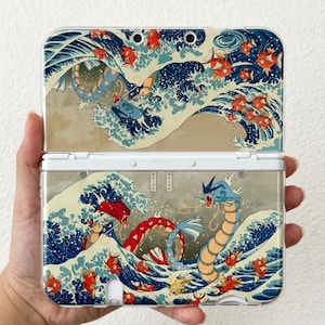 May include: A handheld gaming console featuring a clear protective case with a printed design of blue ocean waves, numerous orange fish, and a large blue sea serpent creature. The artwork incorporates traditional Japanese wave motifs.