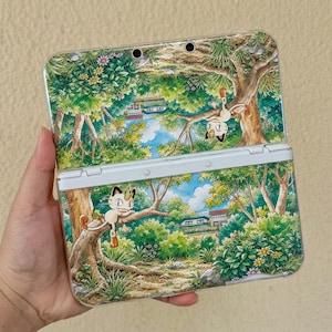 May include: A handheld gaming console featuring a clear protective case decorated with a vibrant forest scene. The artwork displays a cream-colored cat-like creature perched on tree branches amidst lush green foliage, flowers, and a small house.