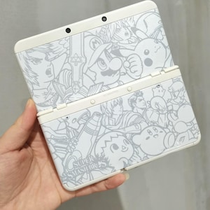 Może przedstawiać: Biała konsola do gier przenośna Nintendo 3DS z szarym wzorem graficznym przedstawiającym postacie z serii gier wideo Super Smash Bros. Konsola jest otwarta, ukazując ekran i przyciski.