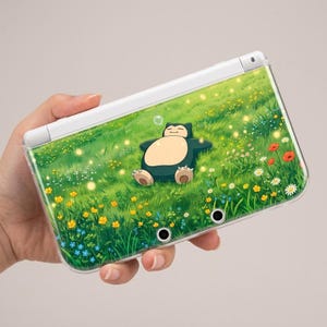 May include: A white handheld gaming console featuring a protective cover with a printed illustration of a Snorlax character lying on its back in a vibrant green meadow filled with yellow, white, and red wildflowers and glowing fireflies.