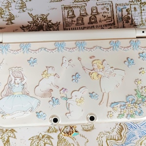 Może przedstawiać: Kremowe etui na Nintendo 3DS z wytłoczonymi ilustracjami dziewczynki, kota, królika i wróżki. Projekt zawiera motyle, kwiaty i kokardy w pastelowych kolorach. Etui ma przezroczystą plastikową górę.