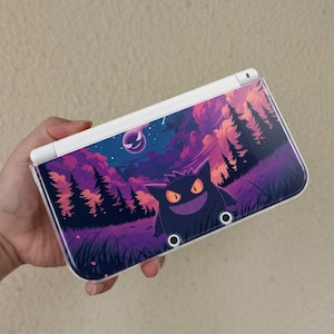 May include: A white handheld gaming console featuring a custom skin with a purple Gengar character in a dark forest landscape. The graphic displays a starry night sky with pink and purple clouds, silhouetted pine trees, and a glowing moon.