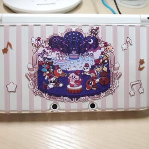 May include: A white and pink striped Nintendo 3DS case featuring a colorful illustration of cartoon characters in a circus-like setting. The design includes musical notes and stars. The case has a clear protective cover.