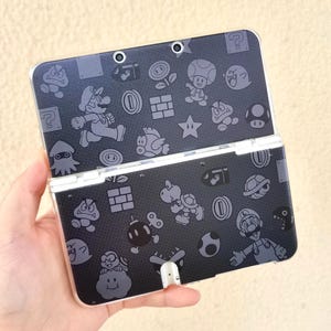 Op de afbeelding: Een zwarte Nintendo 3DS handheld gameconsole met een grijs patroon van Mario-personages en iconen. De console is open, met het scherm en de knoppen zichtbaar. Het ontwerp bevat Mario, munten en andere game-elementen.