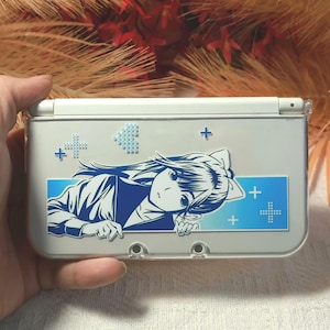 May include: A white Nintendo 3DS handheld game console with a custom design. The design features a blue and white anime-style illustration of a girl, along with blue cross and dot patterns. The console is held in a hand.