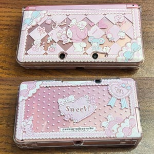 Può includere: Due console Nintendo 3DS rosa con cover decorative. La console superiore presenta un design con un personaggio dei cartoni animati, fiocchi e la scritta "Chu!". La console inferiore ha un motivo a pois con la scritta "Sweet!" in un cuore.