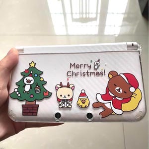May include: A white Nintendo 3DS case featuring Christmas-themed cartoon characters. The case has a Christmas tree with a penguin, a reindeer, a chick, and a bear in a Santa suit. The text "Merry Christmas!" is also visible.