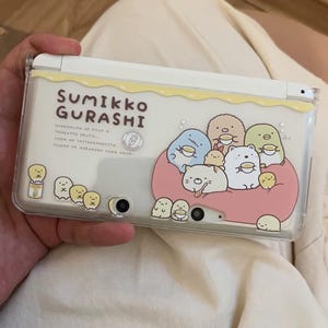 Può includere: Custodia bianca per console di gioco portatile con la scritta "Sumikko Gurashi" e personaggi dei cartoni animati. La custodia ha una copertura in plastica trasparente con un bordo giallo e un design a cuscino rosa.