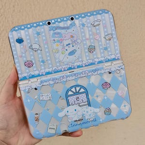 May include: A light blue Nintendo 3DS case with a Cinnamoroll design. The case has a clear plastic overlay with a repeating pattern of white diamonds, bows, and cartoon illustrations. The case is decorated with a variety of cute illustrations, including a book, a cup of boba, and a cupcake.