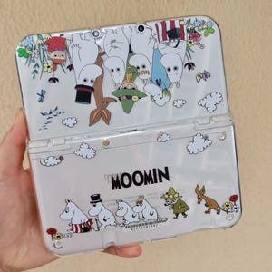 May include: A clear protective case for a handheld gaming console featuring Moomin characters. The design includes Moomin, Snufkin, and Little My among colorful flowers and clouds. The word MOOMIN is printed in black on the lower section of the case.
