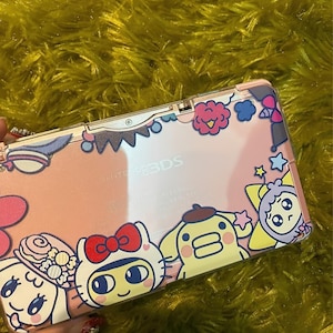 May include: A pink Nintendo 3DS handheld game console with a decorative cover featuring cartoon characters and floral designs. The cover has a pink background with white, blue, yellow, and red accents. The text "Nintendo 3DS" is visible.