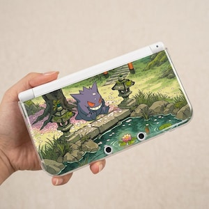 May include: A white handheld gaming console featuring a decorative skin with a purple Gengar character sitting by a stone lantern, a pond with a pink water lily, and cherry blossom petals scattered across a green landscape.