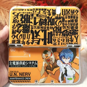 Puede incluir: Una funda naranja y blanca con texto japonés e ilustraciones de estilo anime. La funda tiene un diseño con bisagras e incluye el texto "MAIN ENERGY SUPPLY SYSTEM" y "U.N. NERV MAINTENANCE CREW".