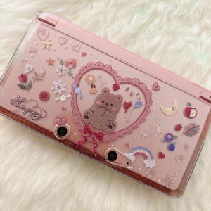 May include: Pink handheld gaming console case with a clear overlay. The case is decorated with a heart-shaped design featuring a bear, flowers, and the word "Happy". Additional embellishments include a rainbow, moon, and stars.