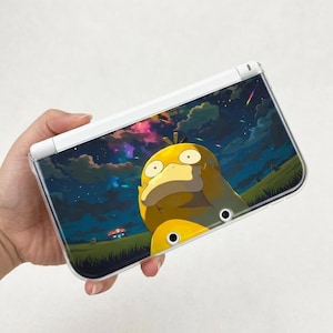 May include: A white handheld gaming console featuring a protective cover with a graphic illustration of a yellow Psyduck character against a dark, starry night sky landscape with clouds and a small building in the distance.
