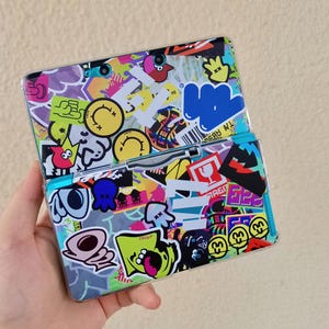 Puede incluir: Una consola de juegos portátil cubierta de pegatinas coloridas. Las pegatinas presentan varios personajes de dibujos animados, formas y texto, incluyendo caras sonrientes, una nube azul y un calamar rojo. La consola está abierta, revelando la pantalla y los botones.
