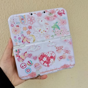 May include: White handheld gaming console case with a floral and bunny design. The case features pink, red, and yellow flowers, a white bunny, butterflies, and a heart shape. The case is open, revealing the screen and buttons.