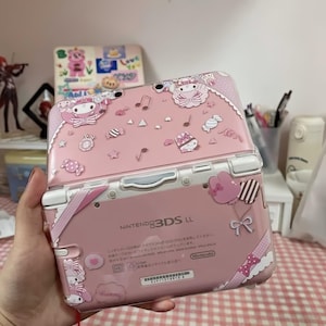 Könnte beinhalten: Rosa Nintendo 3DS LL Handheld-Spielkonsole mit dekorativer Hülle mit Cartoon-Figuren, Noten und Süßigkeiten. Die Konsole ist geöffnet und zeigt den Bildschirm und die Tasten. Die Hülle hat eine transparente Kunststoffhülle.