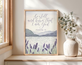 May include: A framed watercolor print featuring the text "Be still and know that I am God" above a landscape of a lake, mountains, and lavender. The print is in a light wood frame and is displayed on a wooden surface.