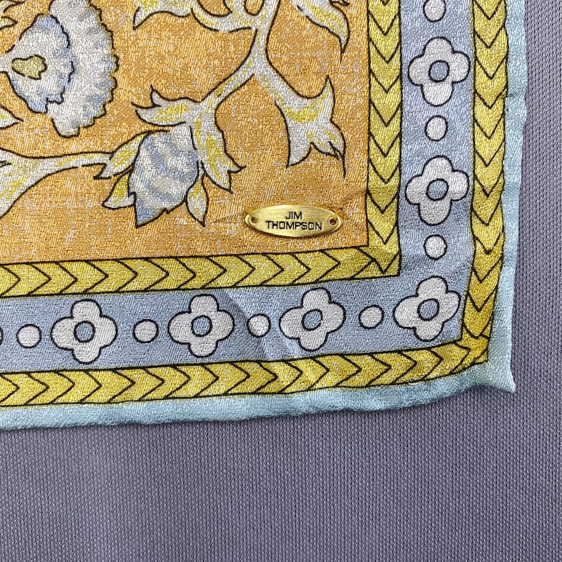 Puede incluir: Un pa&ntilde;uelo de seda cuadrado con un centro amarillo dorado y un patr&oacute;n floral en blanco y azul claro. El borde presenta un dise&ntilde;o de chevr&oacute;n amarillo y una banda azul claro con motivos florales blancos. Una etiqueta de metal dorado dice JIM THOMPSON.