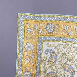 Puede incluir: Un textil cuadrado que presenta un panel central blanco con un patr&oacute;n floral y de cachemira en azul claro y verde, rodeado por un borde amarillo mostaza con motivos florales azules y un ribete azul claro con un dise&ntilde;o de tr&eacute;bol blanco.