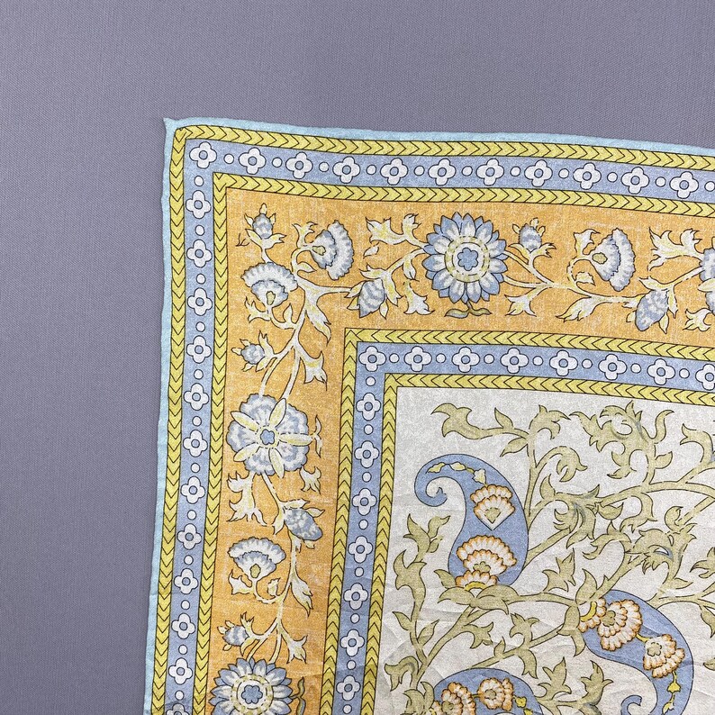Puede incluir: Un textil cuadrado que presenta un panel central blanco con un patr&oacute;n floral y de cachemira en azul claro y verde, rodeado por un borde amarillo mostaza con motivos florales azules y un ribete azul claro con un dise&ntilde;o de tr&eacute;bol blanco.