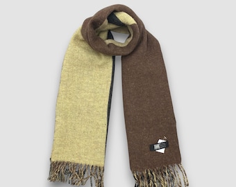 May include: A two-tone scarf in shades of brown and tan with fringed ends. The scarf is folded over, showcasing the contrasting colors. A small tag is attached to the scarf.