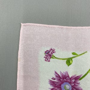 Puede incluir: Un textil cuadrado rosa claro con un dise&ntilde;o floral. El dise&ntilde;o presenta flores moradas con tallos y hojas verdes. El fondo es de color rosa claro.