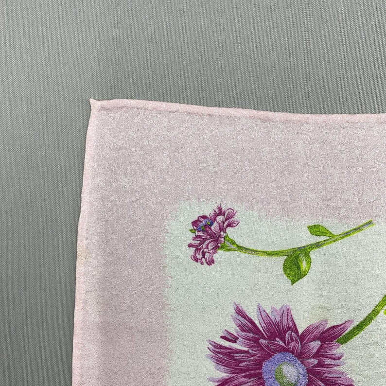Puede incluir: Un textil cuadrado rosa claro con un dise&ntilde;o floral. El dise&ntilde;o presenta flores moradas con tallos y hojas verdes. El fondo es de color rosa claro.